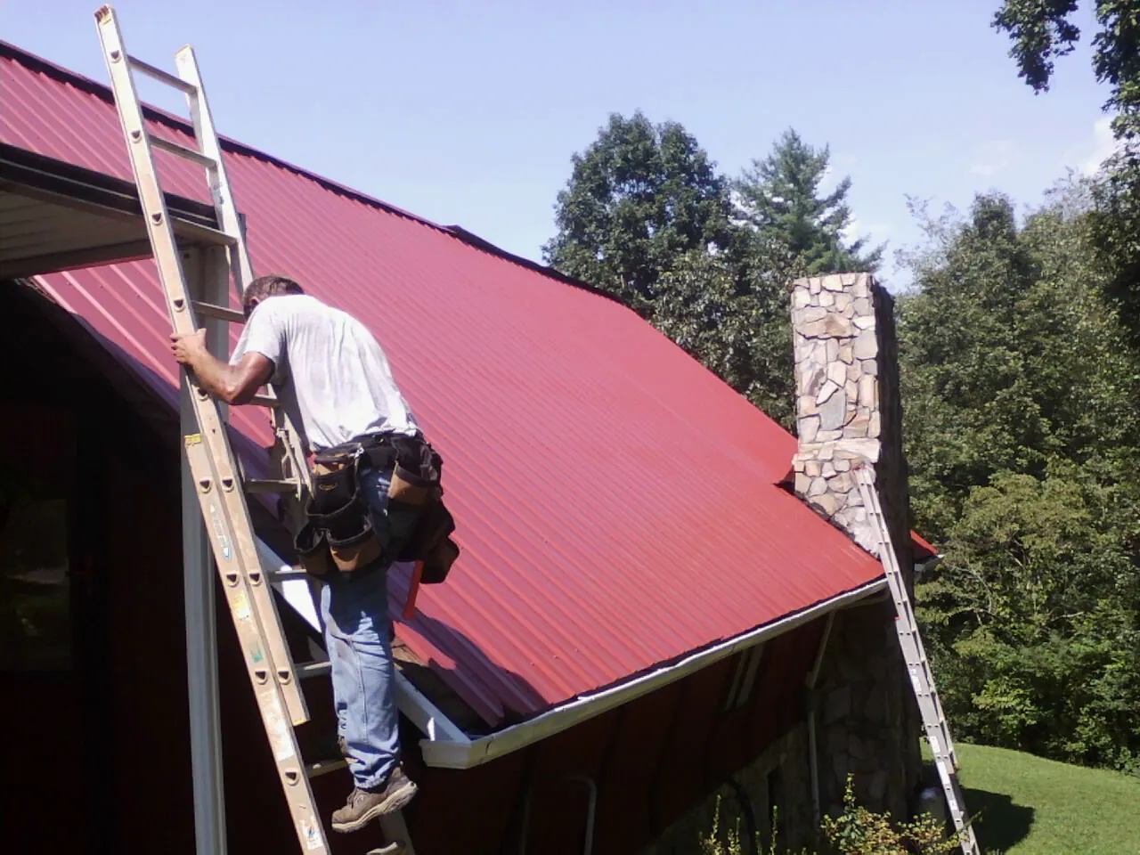 Roofer on a ladder beside a red metal roof and stone chimney — Soffit & Fascia Repair by Roofing Brothers in Tetonia